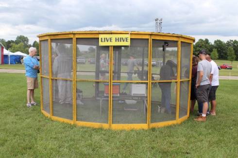 Photo of people standing outside of a screened tent with a honey bee hive inside the tent, sign reads 