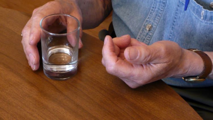 A shot of an elderly person's hands as they take medication, with a pill in one hand and a glass of water in the other.