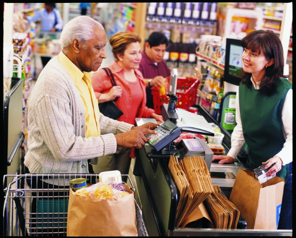 A man using the payment keypad at a grocery store checkout while a clerk bags the groceries.