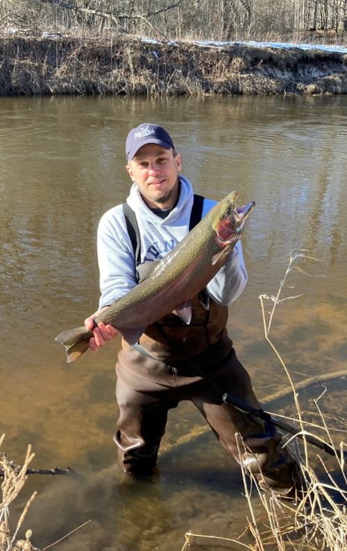 Man wearing waders is seen standing in the river holding a steelhead he caught.