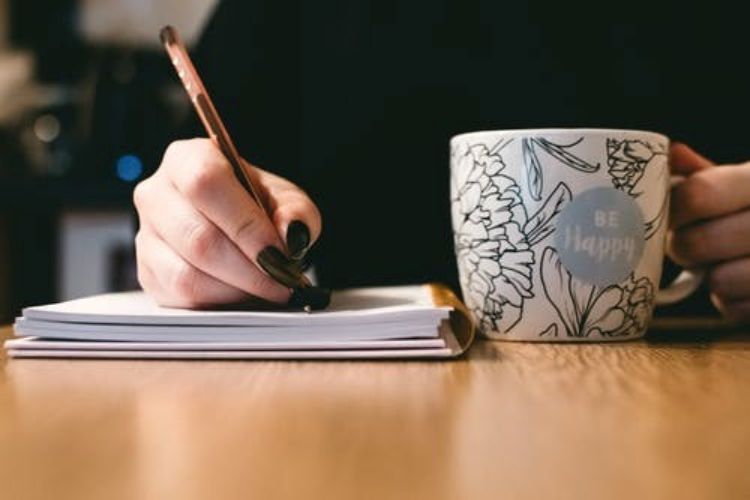 Closeup of person's hands as they write in a notebook, with a pencil in one hand and a coffee mug in the other.