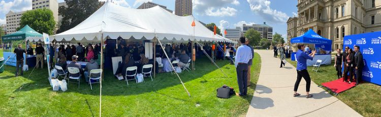 A large white tent is shown from a distance with people inside at a trade show event outside the Michigan capital on a very sunny day.