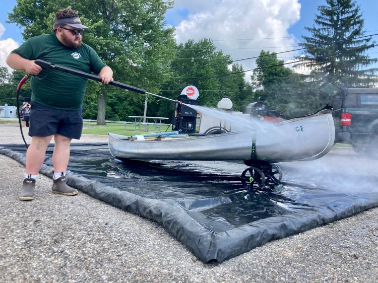 Man using hot water, high pressure spray washer to clean metal canoe in parking lot.