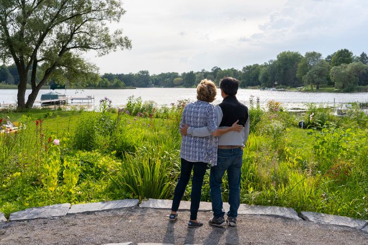 A man and woman with arms wrapped around each other looking out at a lake.