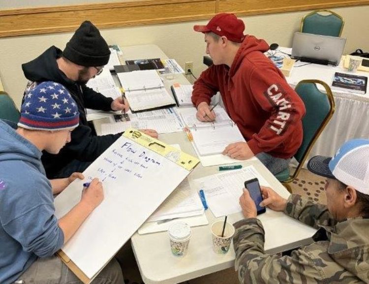 Four participants are gathered around a table reviewing paperwork and one is writing on a large paper tablet as they review a case study in their training class. Photo: Laurie White, GLIFWC