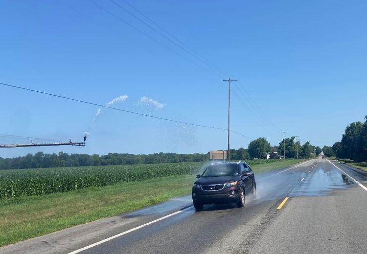 A car driving on the road gets soaked by water shooting out of an irrigation system that's in the field next to the road.
