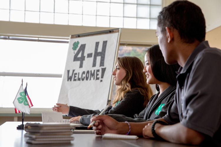 Three youth sitting at a table next to a sign that says 4-H Welcome.