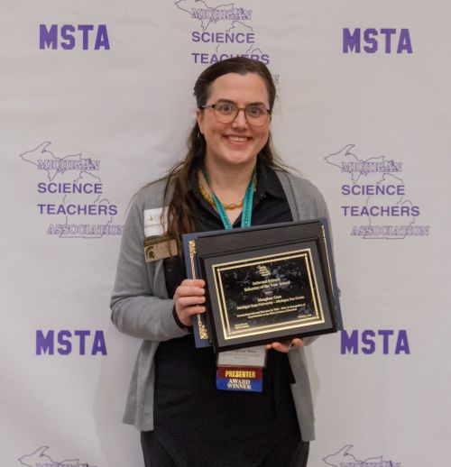 Woman stands in front of a banner that says MSTA, holding an award  plaque.