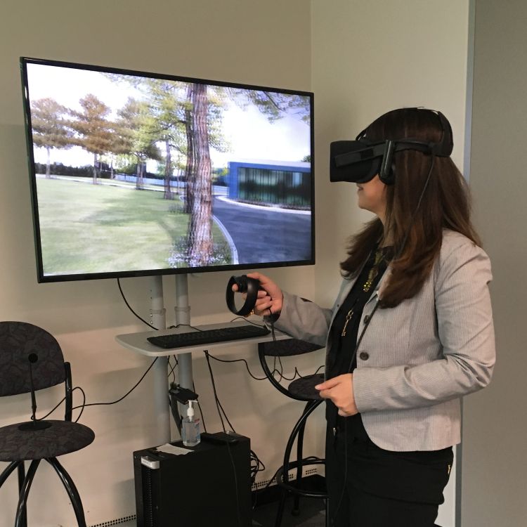 A woman wearing virtual reality head gear demonstrates use of this equipment with hand-held controls with a monitor in front of her showing her changing perspectives on the screen.
