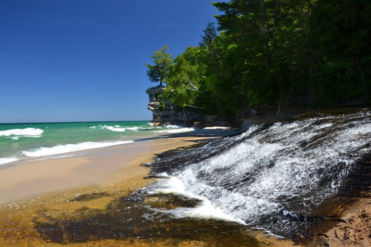 A view of Lake Superior shoreline that includes a sandy beach, a waterfall and the cliffs of Pictured Rocks in the background.