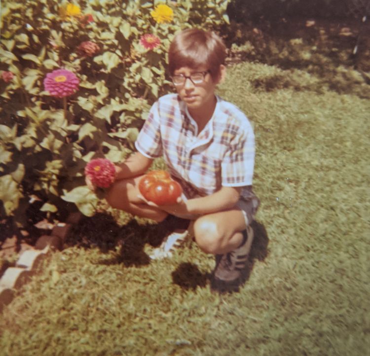 Photo of David Douches, aged 12,  kneeling next to his flower garden with tomato in hand from 1972