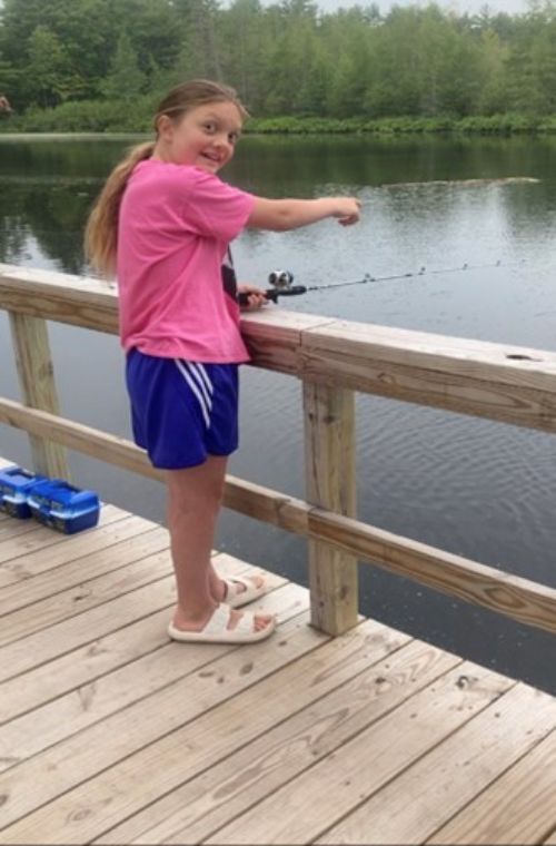 A young girl stands on a fishing pier with her fishing pole. She is looking over her right shoulder toward the camera and smiling.