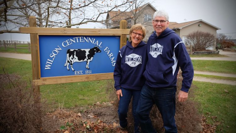 Nancy and Brent Wilson standing in front of the Wilson Centennial Farms sign and their farm house.