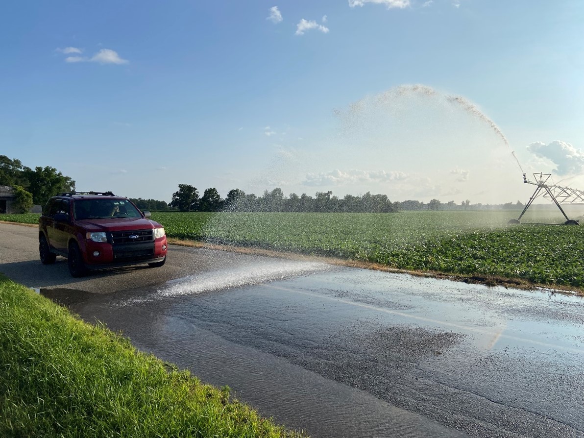 A car on the road stops just short of a line of water from a nearby irrigator shooting into the road.