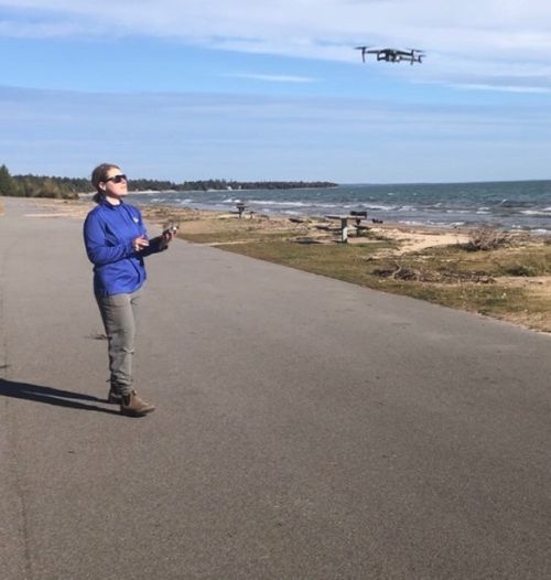 Woman stands near a lakeshore beach and holds drone controls in her hand as she lands the drone that can be seen in the air near her.