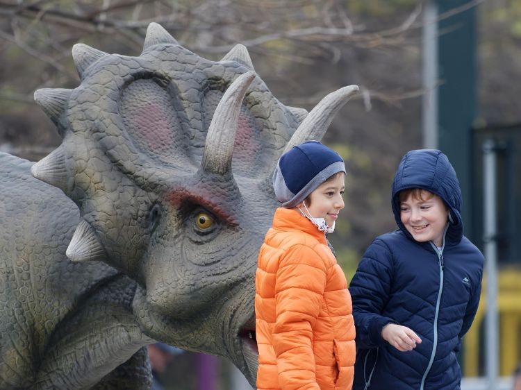 Two young children, one in an orange coat and one in a blue coat, standing outside a large dinosaur replica.