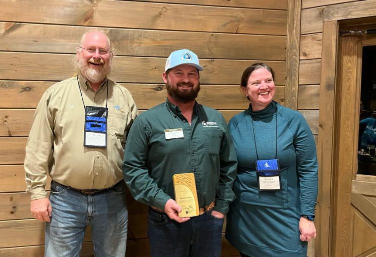 Three people with name tags posing for the camera and smiling with the middle person holding an award