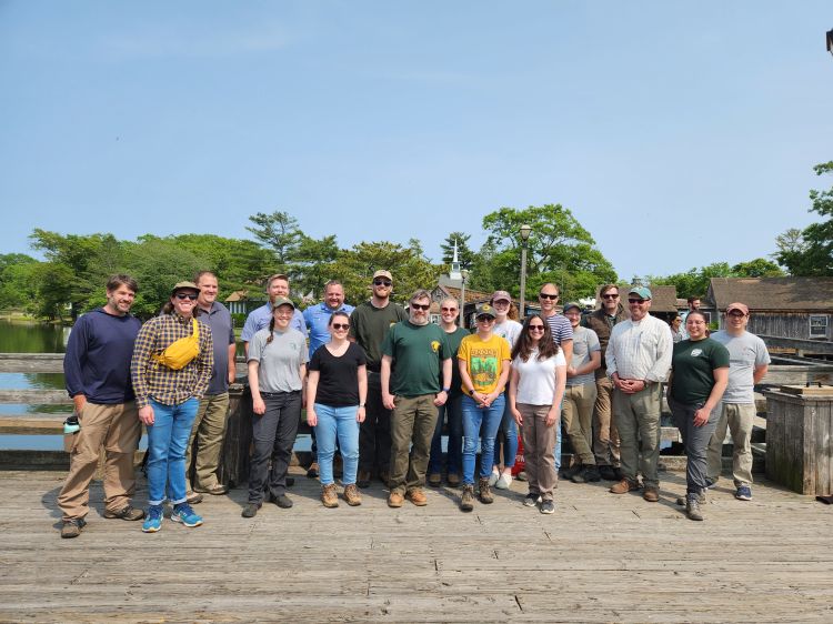 A group of people posing together on a wooden dock with a lake and forest in the background.