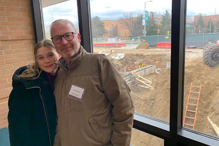 Lily Kirkman with her father Bob Kirkman standing in front of a window. In the background, construction is taking place to renovate the School of Packaging building.