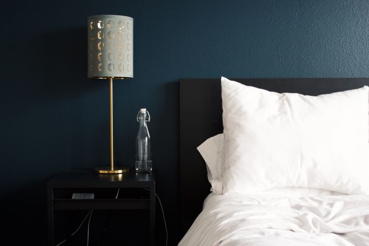 A bed pushed up against a blue wall with a lamp and glass of water on the night stand.