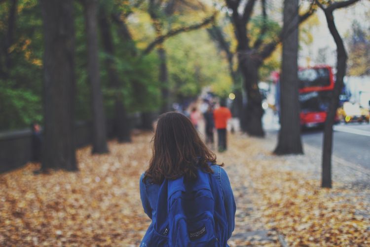 Girl with backpack on walking on sidewalk covered in leaves.