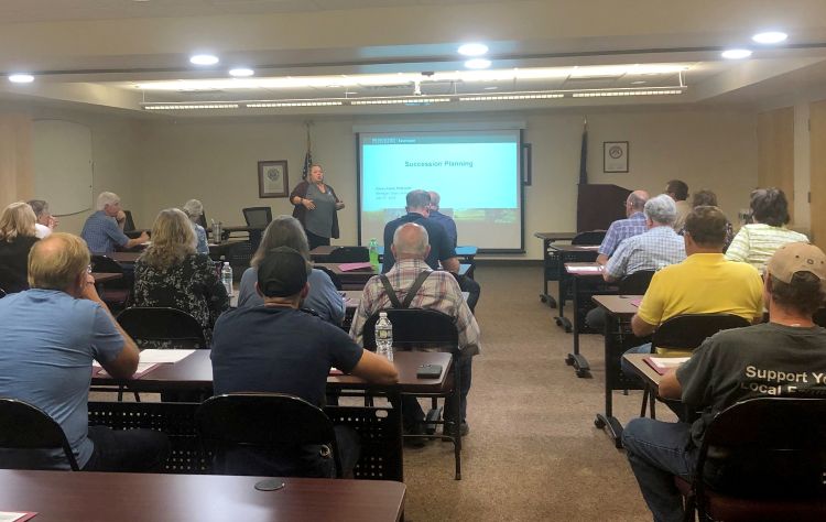 People sitting at desks in a classroom looking up a presenter at the front of the room, standing next to a projector screen.
