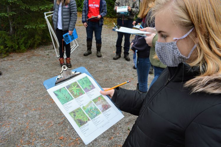 A girl wearing a face mask covering her mouth and nose, holds a clipboard with pictures of plants on it. Other students are seen in the background.