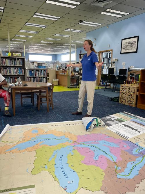 Woman is standing in a library near a large map on the floor of the Great Lakes watershed.