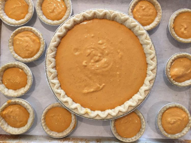 A unbaked pumpkin pie on a sheet surrounded by many mini unbaked pies.