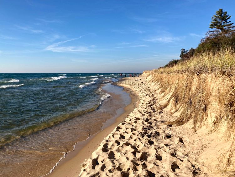 The view looking down the beach on one of the Great Lakes with the water and waves on the left side and a sandy beach rising to a dune on the right.