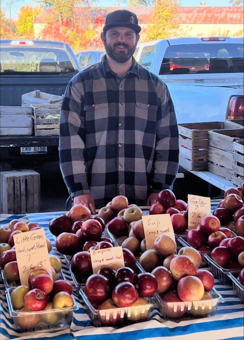 A man standing behind a table at a farmer's market. Apples in containers sit on the table.