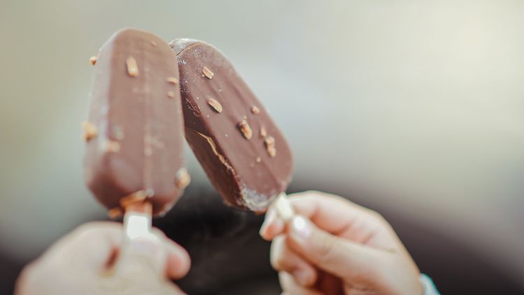 Two vanilla ice cream treats with a chocolate coating being held by two hands leaning toward each other on popsicle sticks.