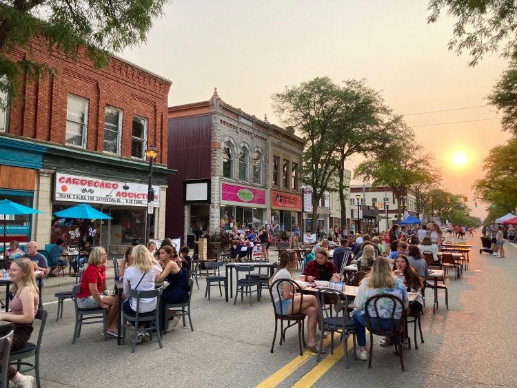 Picture of downtown Alma, Michigan, with restaurant tables set up with people sitting at them on a closed off city street in the evening.