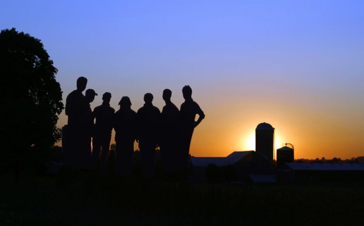 A silhouette of people standing on a farm, with the sun setting in the background over a barn.