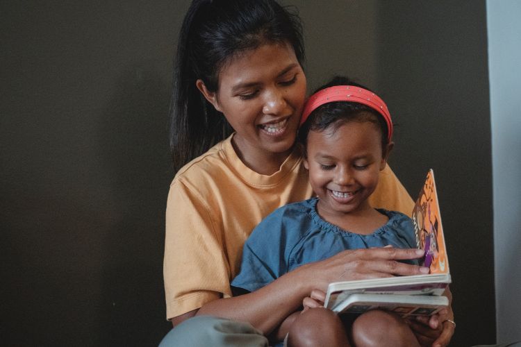 A little girl reads while a woman wraps her arms around the little girl.