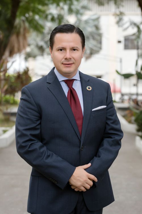 A man in a dark suit and maroon tie with his hands folded in a portrait style stance