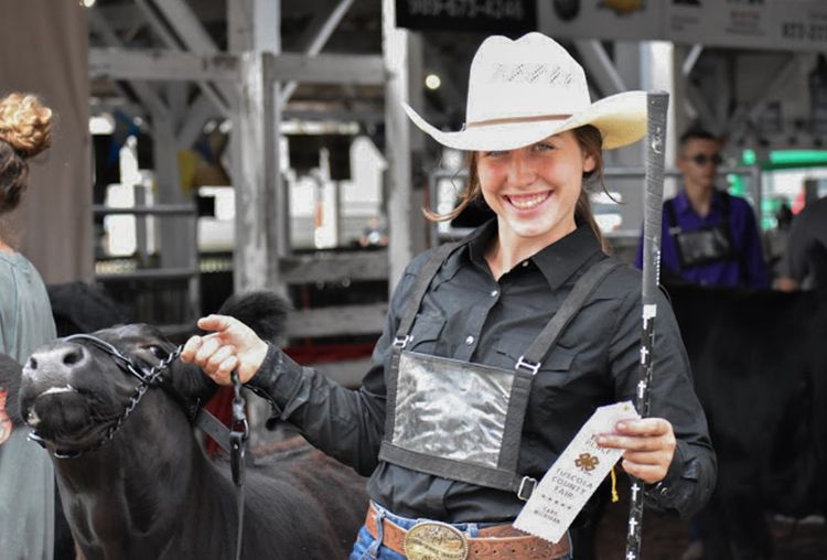 A smiling young woman in a black shirt and white cowboy hat leading a black calf and holding a ribbon.
