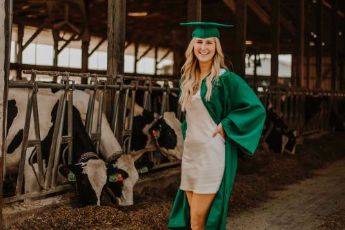 Mikayla Bowen wearing a green graduation cap and gown with MSU dairy cows and a barn in the background.