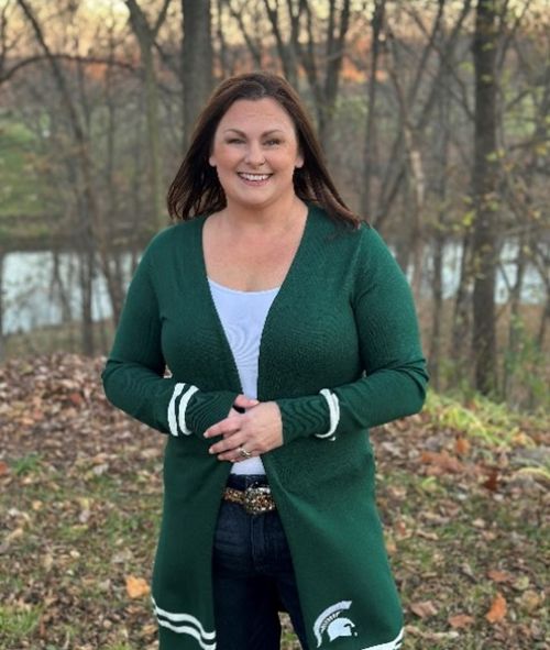 A dark haired smiling white woman in a dark green MSU sweater standing outside.
