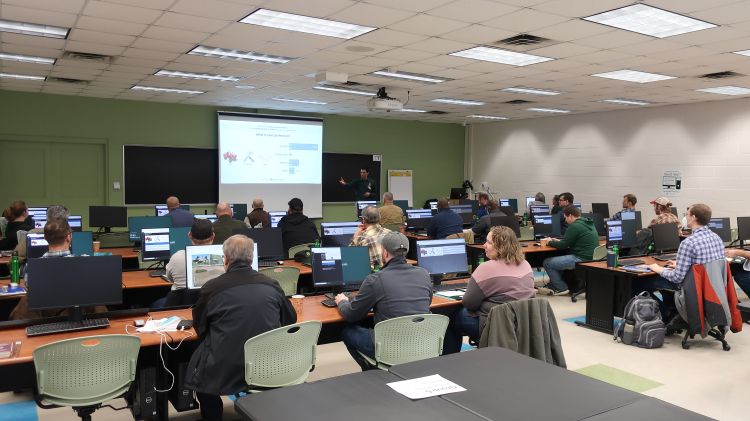 Participants siting at desks with computers in a classroom watching a presenter give a presentation.