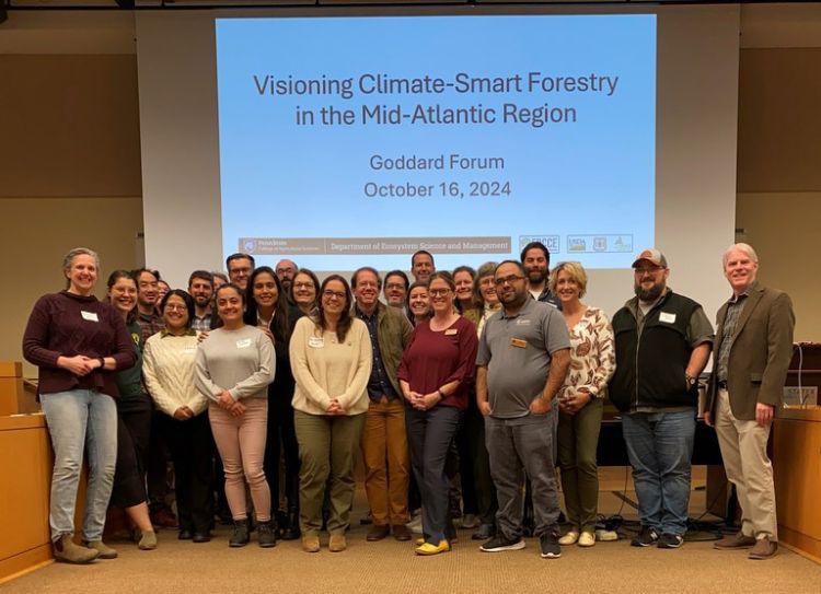 A group photo of participants of the 2024 Goddard Forum standing in front of a projection screen displaying 
