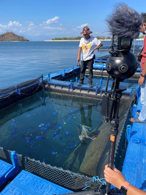 A man stands on the edge of a cage immersed in the water at an ornamental research farm using cage culture aquaculture. In the foreground is someone holding a 360 virtual camera.