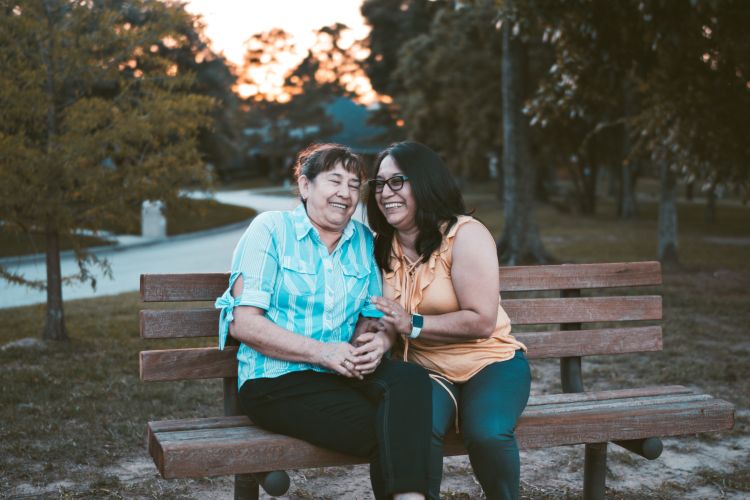 An adult daughter and elderly mother laughing on a park bench together.
