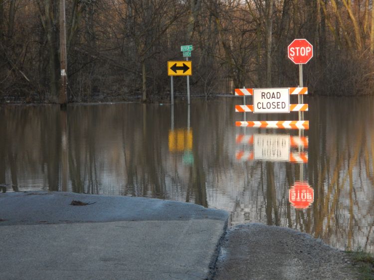 Water is shown covering a road. Road closed sign warns drivers not to enter the roadway.
