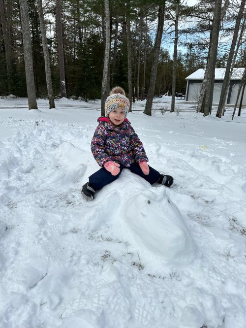 A young girl in black snowpants and a pink and black winter jacket with pink gloves and a hot on top of an alligator form made out of snow.