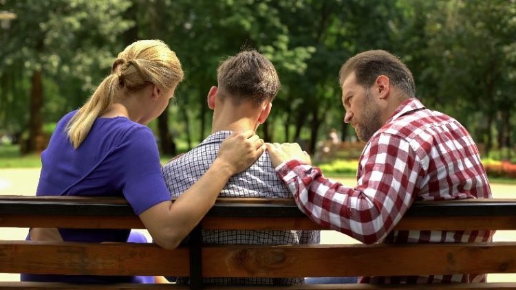 Two adults consoling a younger man on a park bench.