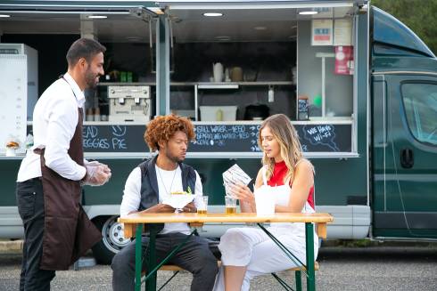 Food truck owner speaking with two customers sitting at picnic table in front of food truck.
