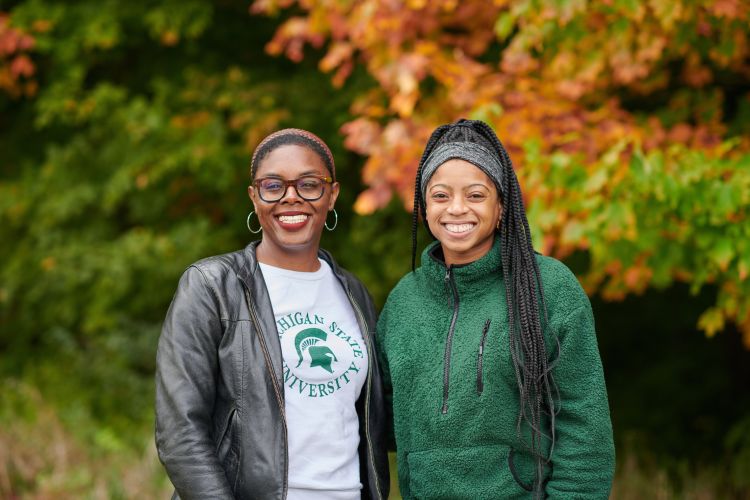 Two women wear Michigan State University shirts standing in front of fall-colored leaves on the edge of a forest.