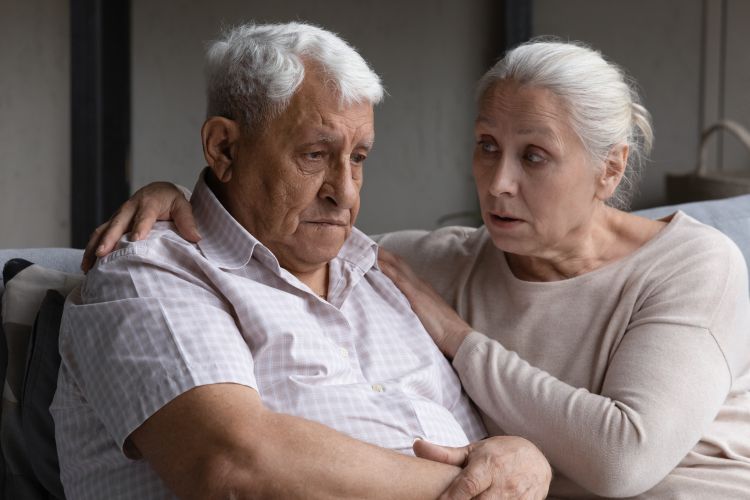 An older woman consoling an older man on a couch.