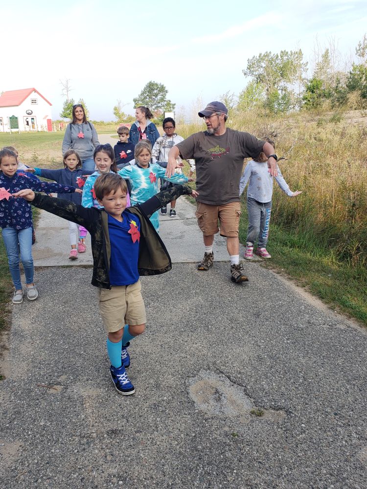 A group of young schoolchildren and their teachers are walking down a sidewalk at Tawas State Park flapping their arms pretending to be birds migrating to the park.